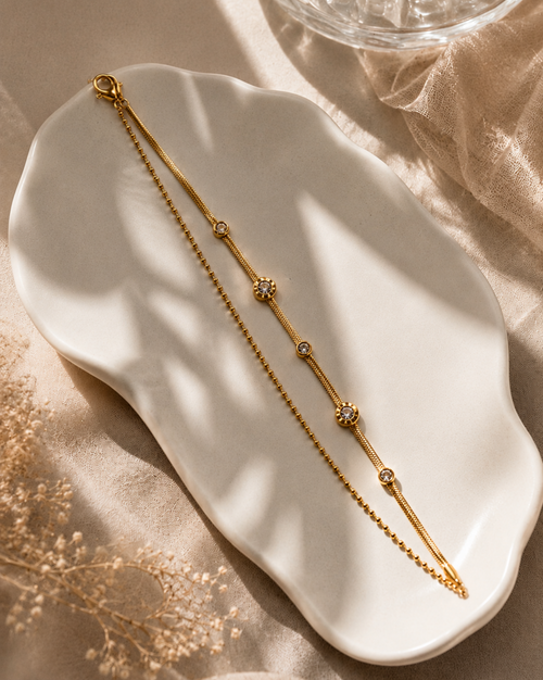 A delicate gold chain bracelet with small floral and oval bead details, displayed on a white ceramic dish over beige fabric with soft shadows.