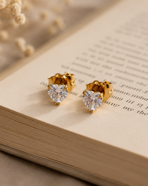 A pair of gold heart-shaped stud earrings with clear heart-shaped gemstones, resting on the pages of an open book with dried flowers in the background.