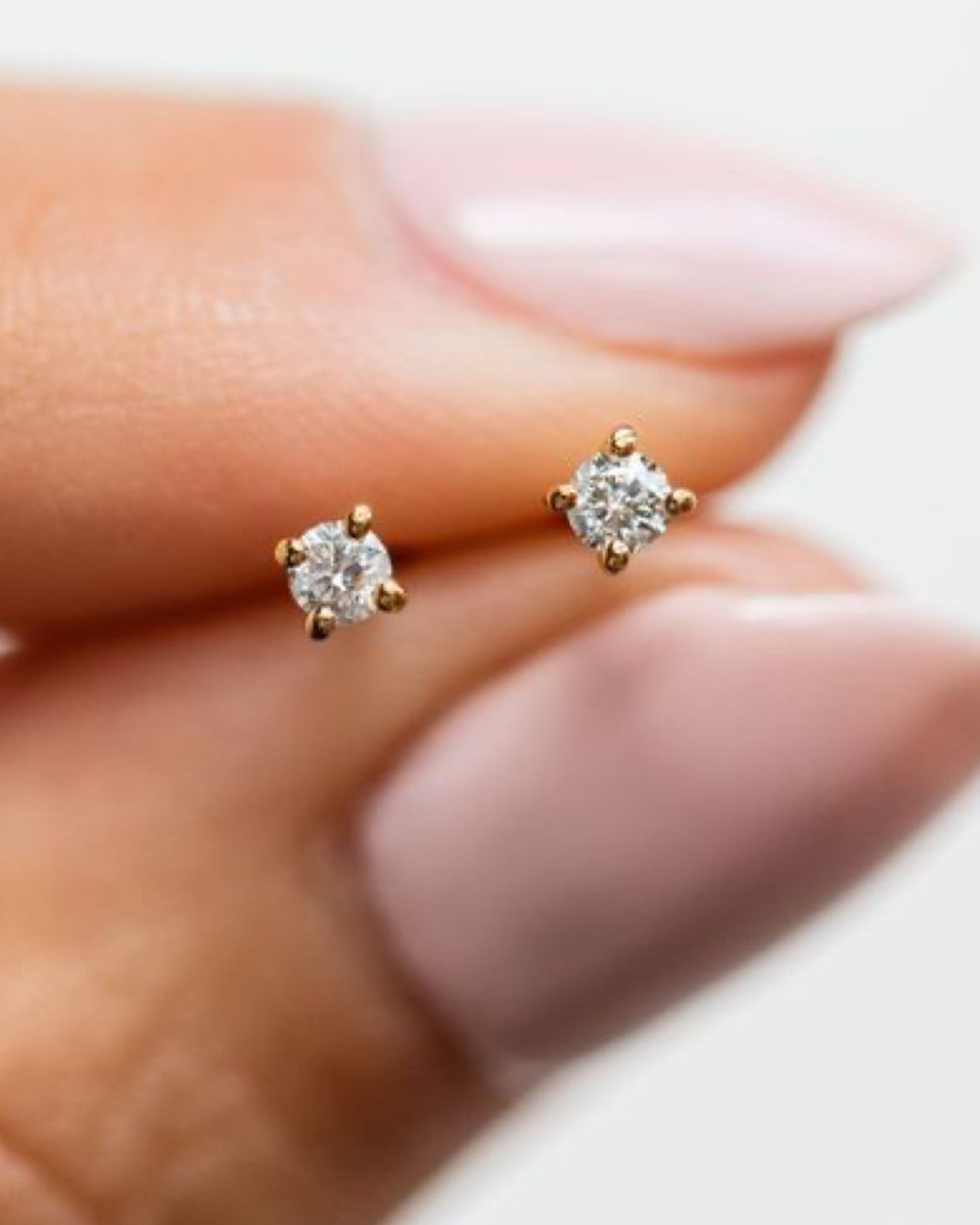 A close-up of a person's hand with pale pink manicured nails wearing a pair of small gold stud earrings with clear round-cut gemstones.