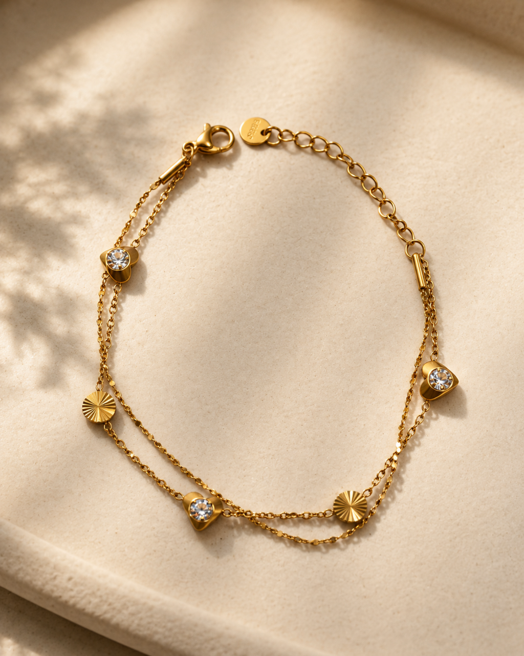 A delicate gold double-strand chain bracelet with small sunburst and heart-shaped charms, some adorned with clear stones, displayed on a soft beige fabric background.