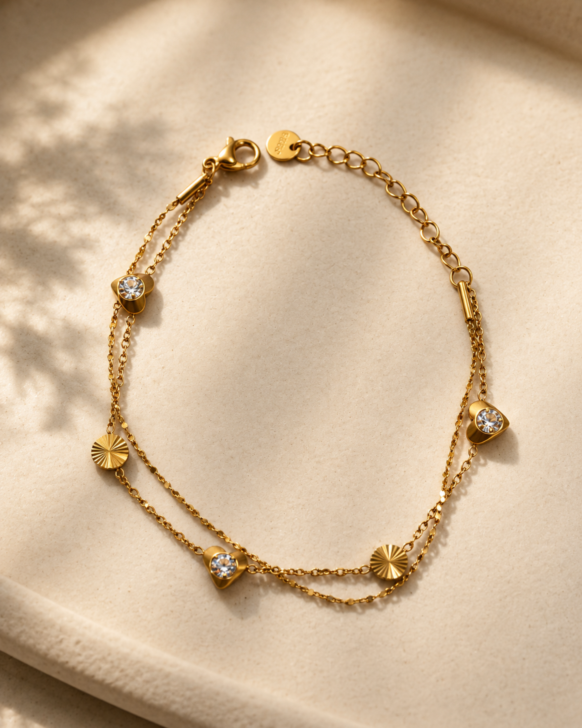 A delicate gold double-strand chain bracelet with small sunburst and heart-shaped charms, some adorned with clear stones, displayed on a soft beige fabric background.