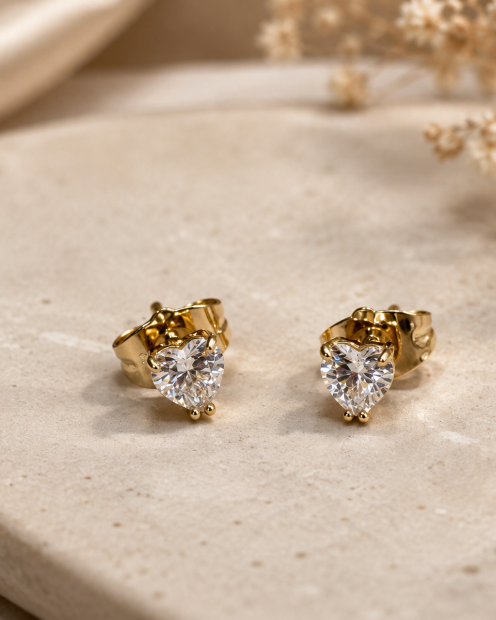 A pair of gold heart-shaped stud earrings with clear heart-shaped gemstones, displayed on a beige stone slab with dried flowers in the background.