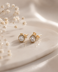 A pair of gold stud earrings with round clear gemstones, displayed on a glossy cream surface next to delicate white baby's breath flowers.