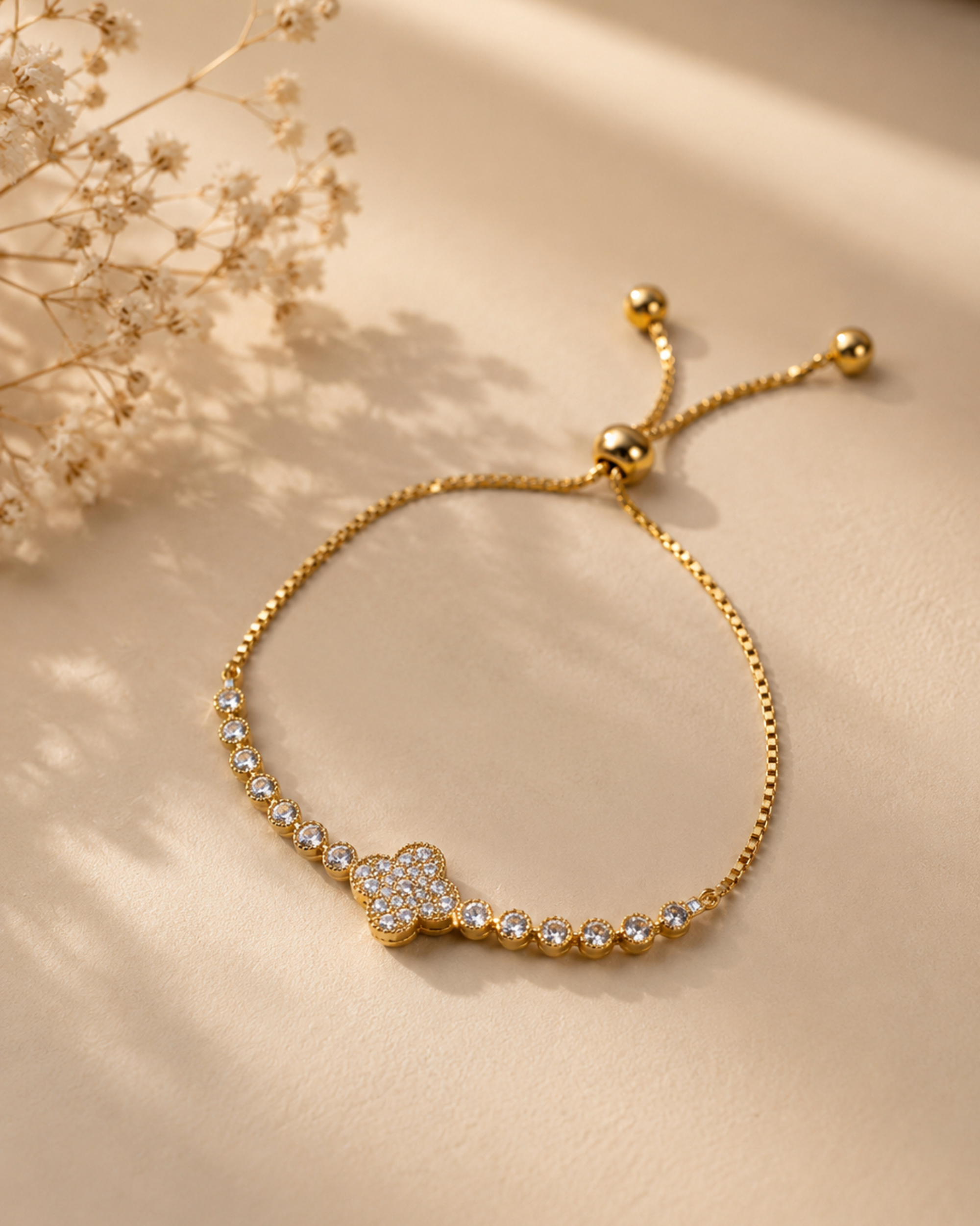 A delicate gold chain bracelet with a central four-leaf clover motif paved with small clear stones, displayed on a warm beige surface with dried floral decor in the corner.