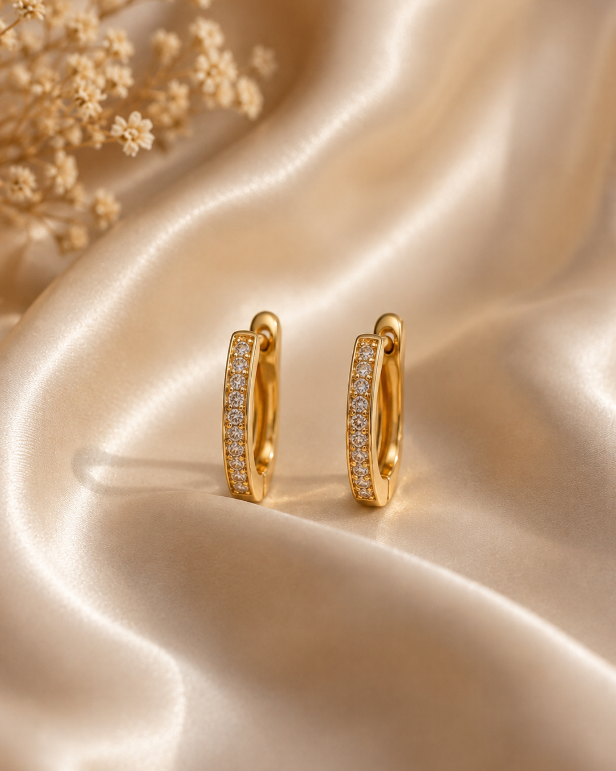 A pair of gold huggie hoop earrings with pavé-set clear stones, displayed on draped champagne satin fabric with dried flowers in the corner.