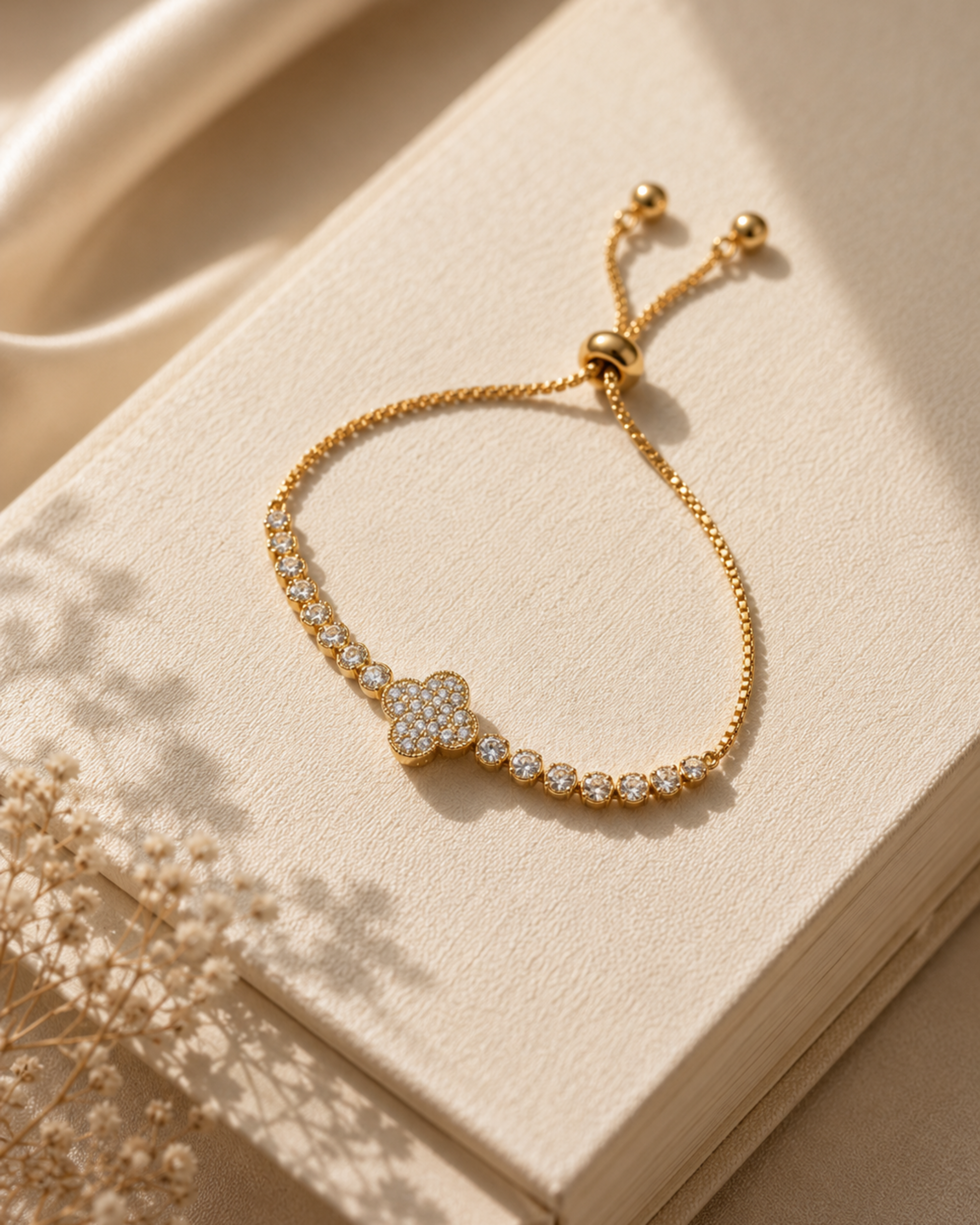 A gold adjustable chain bracelet with a pavé four-leaf clover centerpiece and small round accent stones, displayed on a cream-colored textured book with soft shadows.