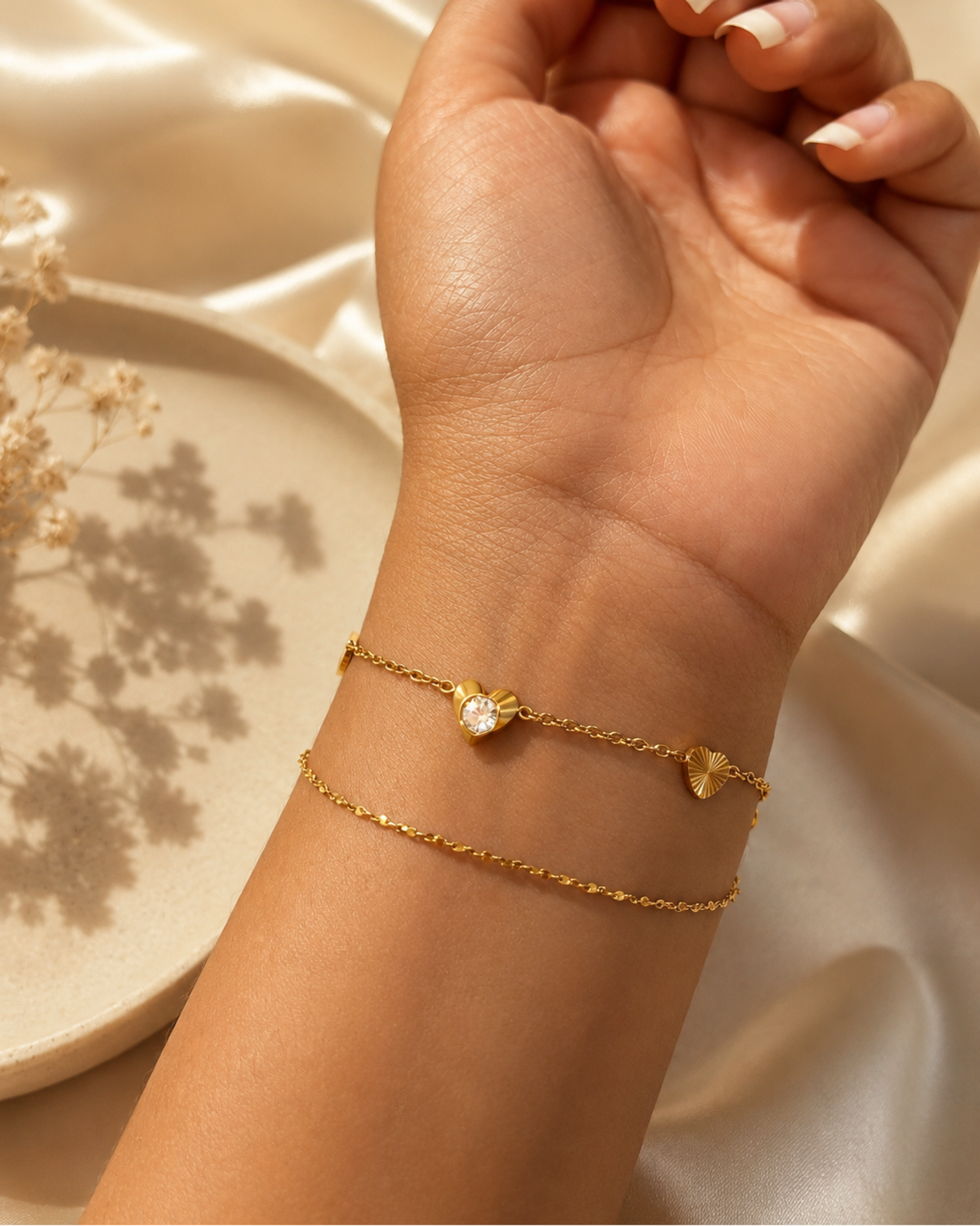 A close-up of a person's wrist wearing two delicate gold chain bracelets, one with a small heart-shaped charm and the other with a small oval charm, set against a soft beige satin background.
