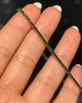 A close-up of a hand with white manicured fingernails holding a delicate tennis bracelet. The bracelet is composed of small, round-cut green gemstones set in a yellow gold-tone metal, creating a vibrant green and gold striped effect.