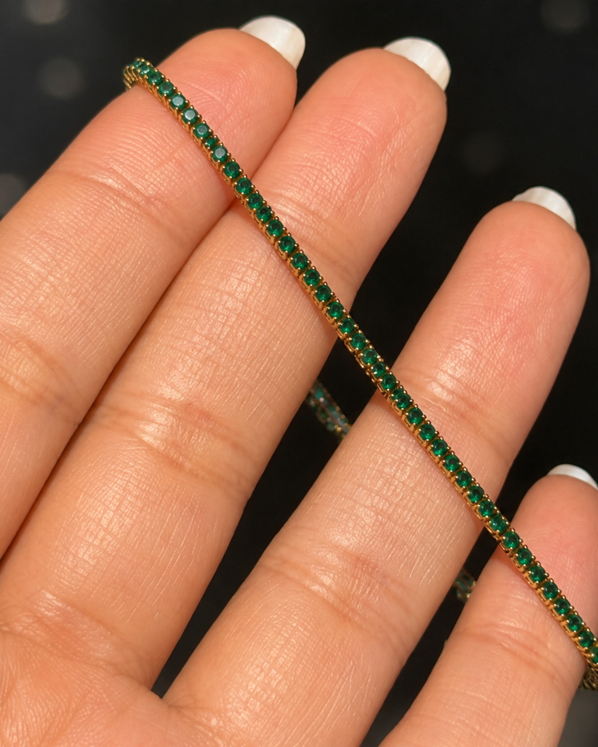 A close-up of a hand with white manicured fingernails holding a delicate tennis bracelet. The bracelet is composed of small, round-cut green gemstones set in a yellow gold-tone metal, creating a vibrant green and gold striped effect.