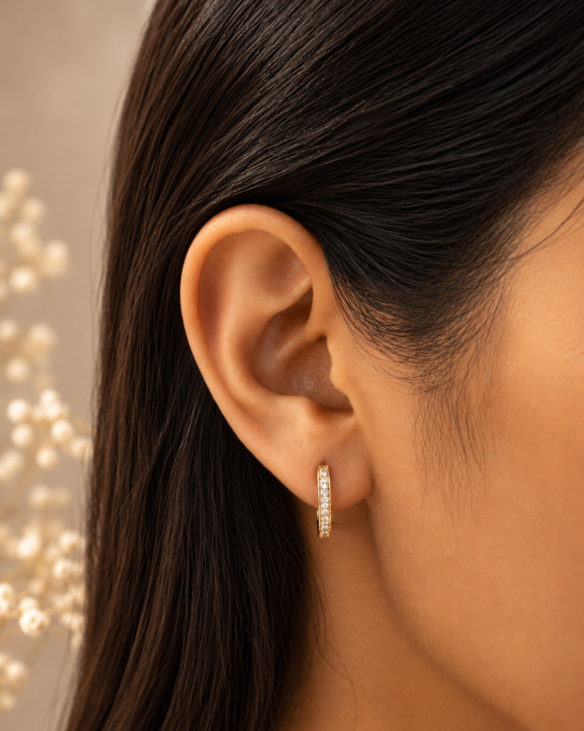 A close-up of a model's ear wearing a small gold huggie hoop earring adorned with clear pavé stones, set against a soft beige background with blurred decorative elements.