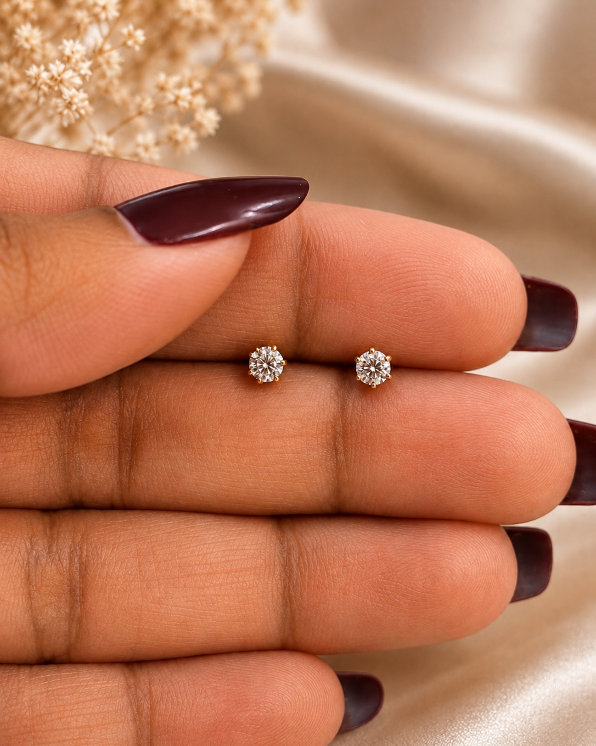 A close-up of a hand with dark burgundy manicured nails holding a pair of small round stud earrings with clear gemstones in a gold-tone setting, against a soft beige satin background with dried flowers.