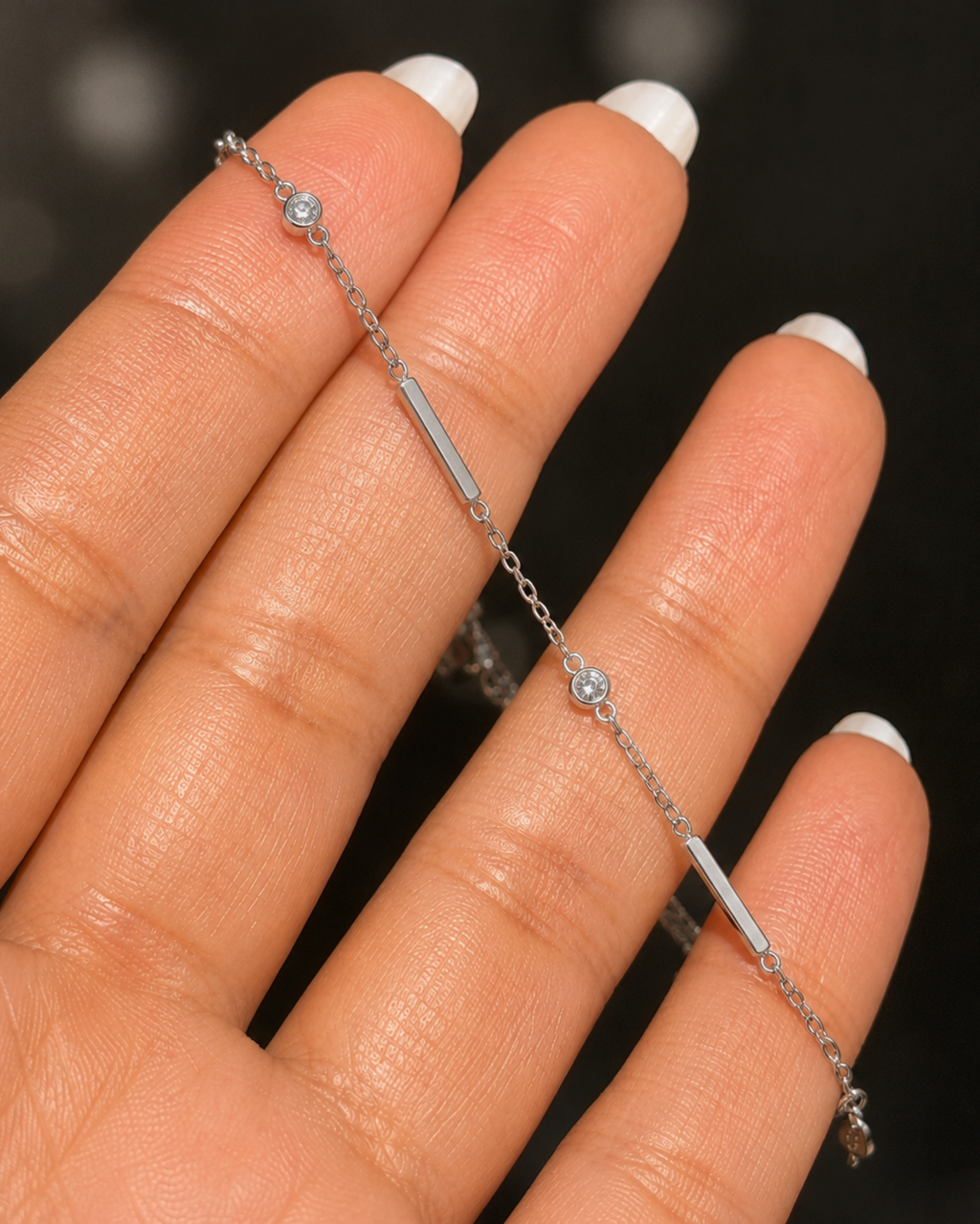 A delicate silver chain bracelet with a central polished bar and small round crystal accents, shown resting on a person's hand with white manicured fingernails against a dark blurred background.