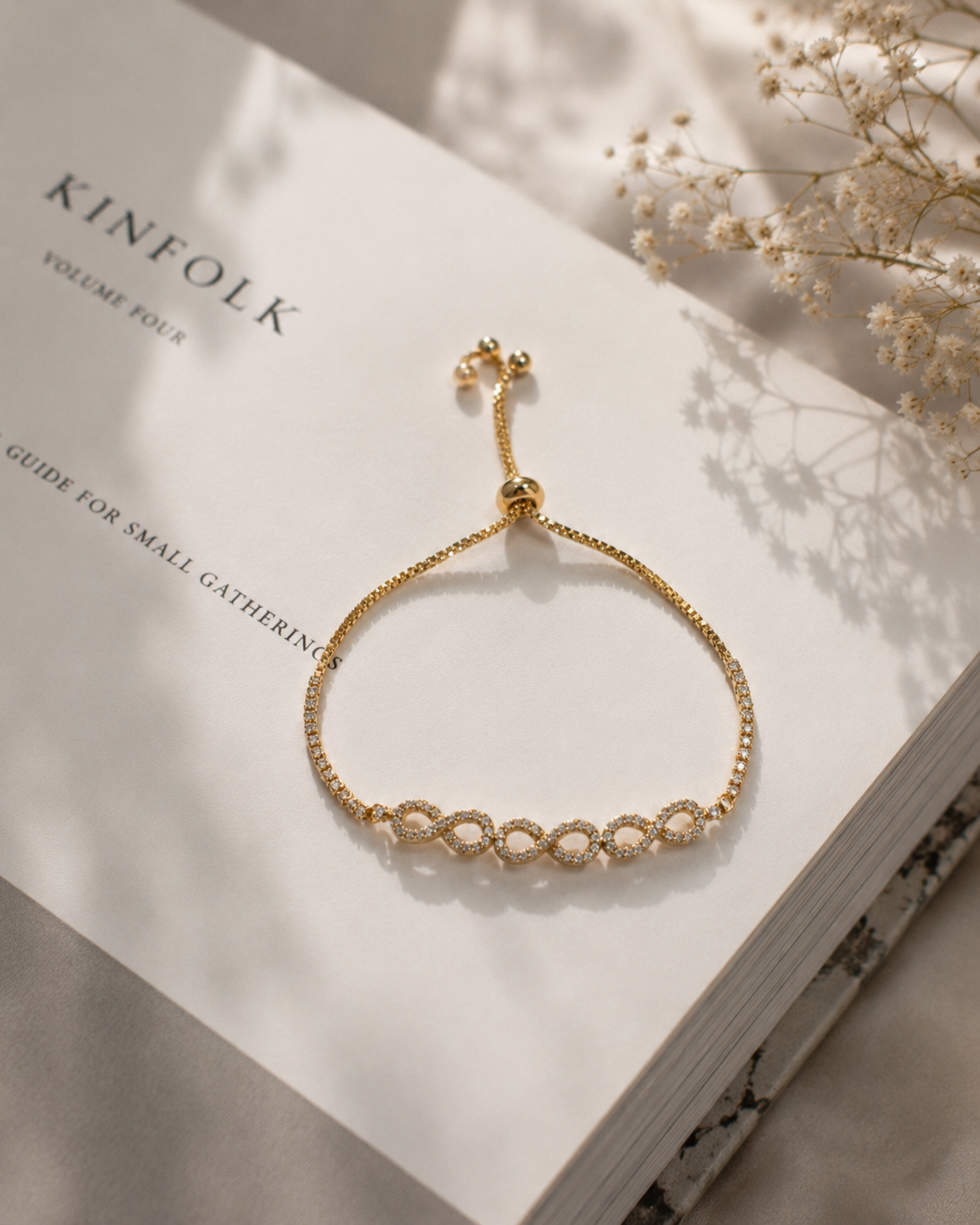 A delicate gold chain bracelet with a row of small clear crystal links, displayed on an open book with dried baby's breath flowers nearby.