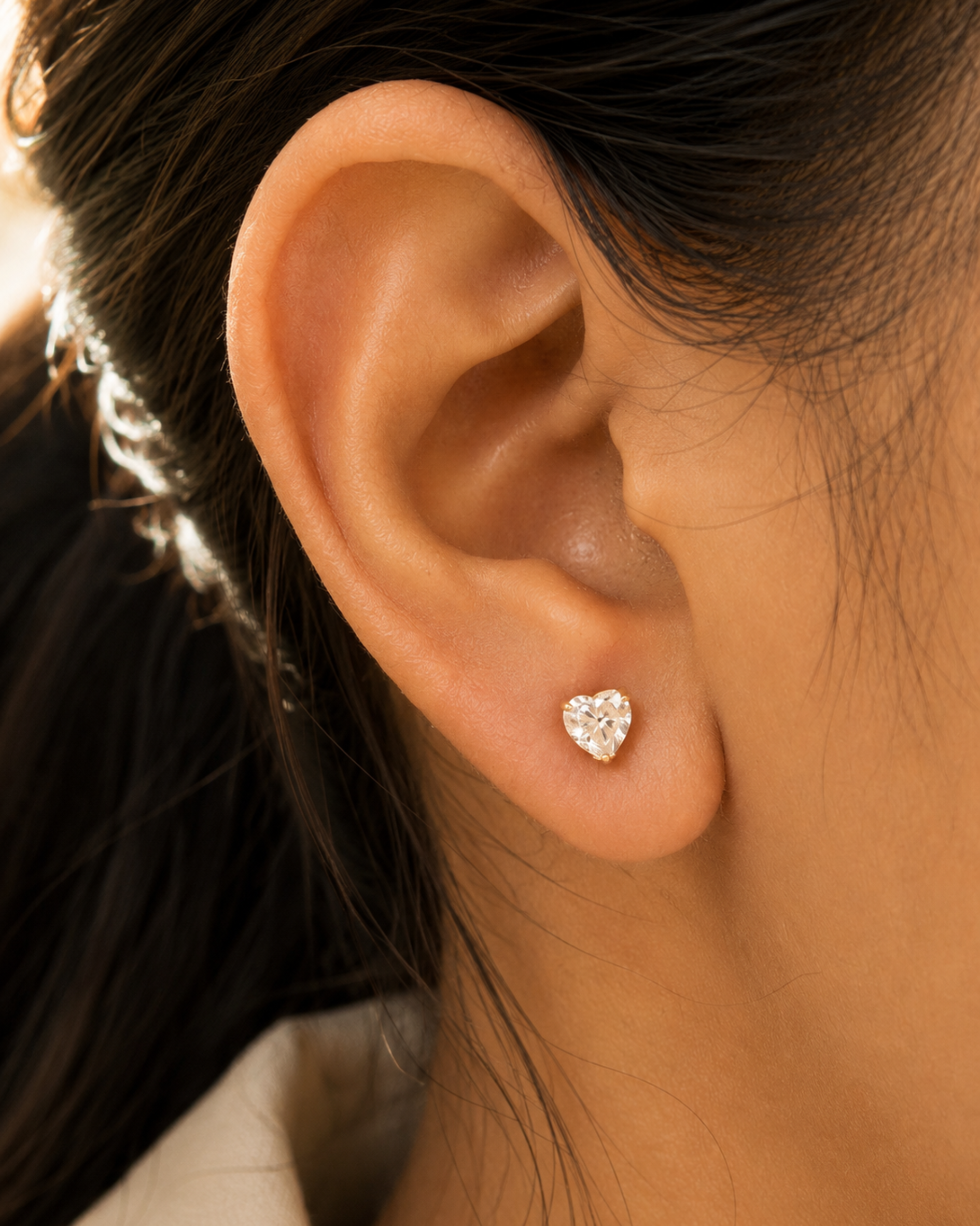 A close-up of a person's ear wearing a small heart-shaped stud earring with a clear sparkling gemstone.