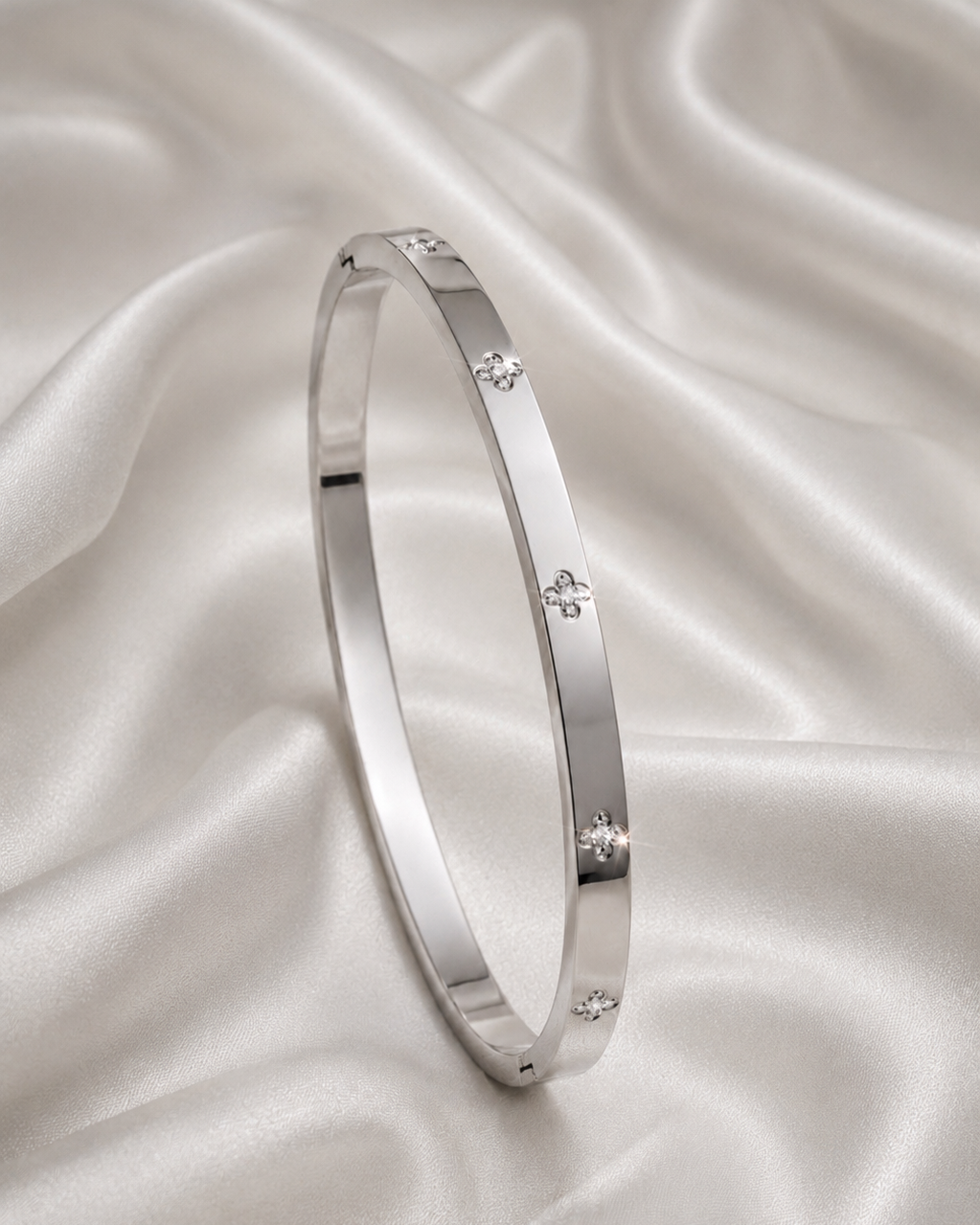 A polished silver bangle bracelet with small diamond accents, displayed on a draped white satin fabric background.