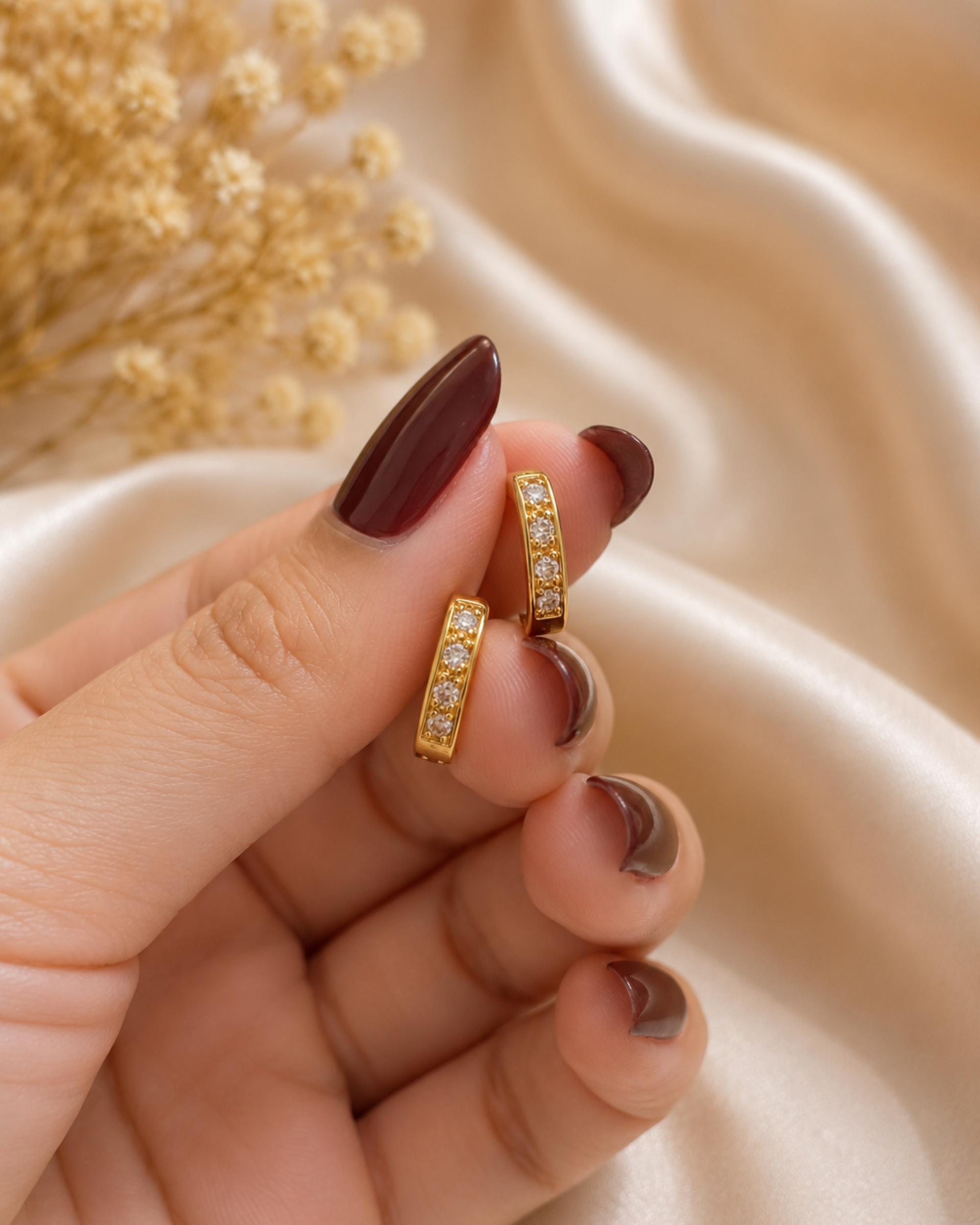 A close-up of a hand with dark brown manicured nails holding a pair of gold hoop earrings adorned with small clear stones, set against a soft beige satin fabric background with dried flowers.