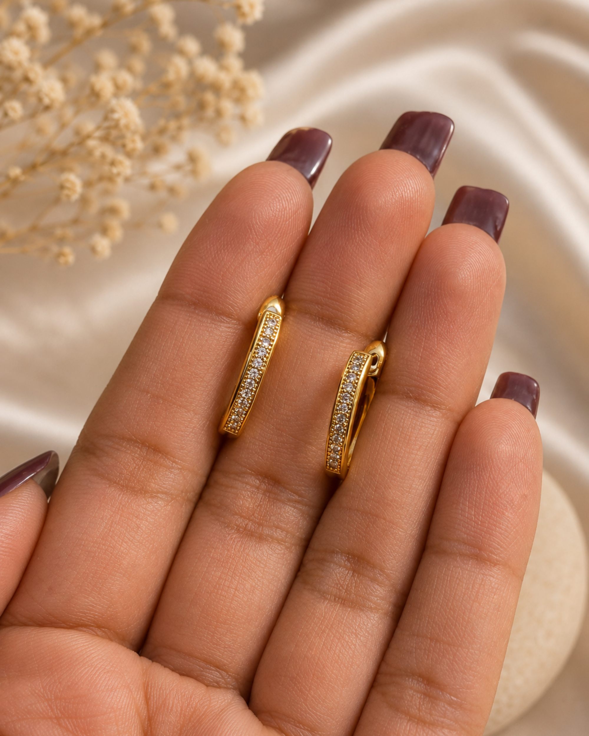 A close-up of a hand with dark plum nail polish holding two small gold huggie hoop earrings with pavé-set clear stones, against a soft cream satin background with dried flowers.