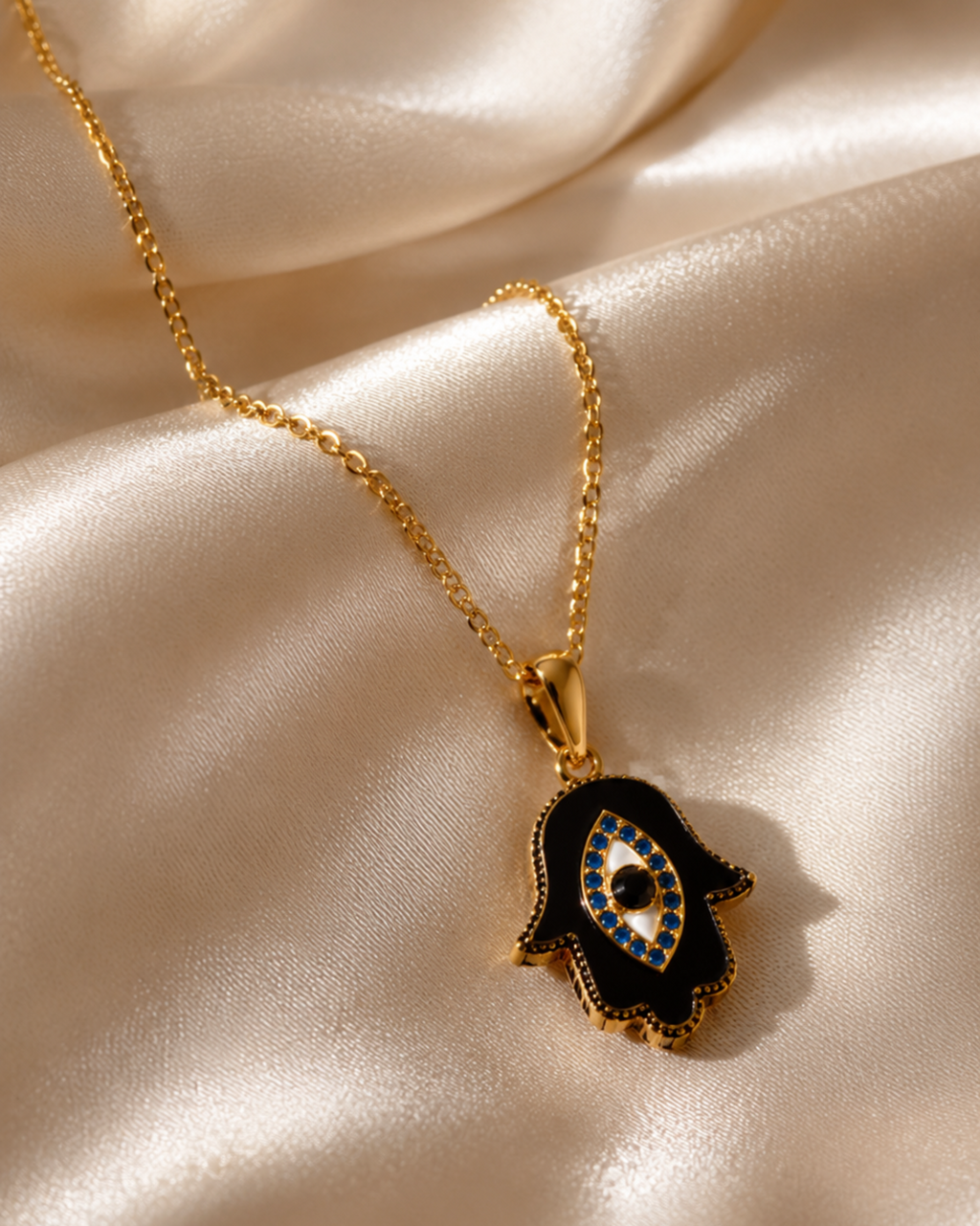 Gold necklace with a black and blue hamsa hand pendant on a beige satin background