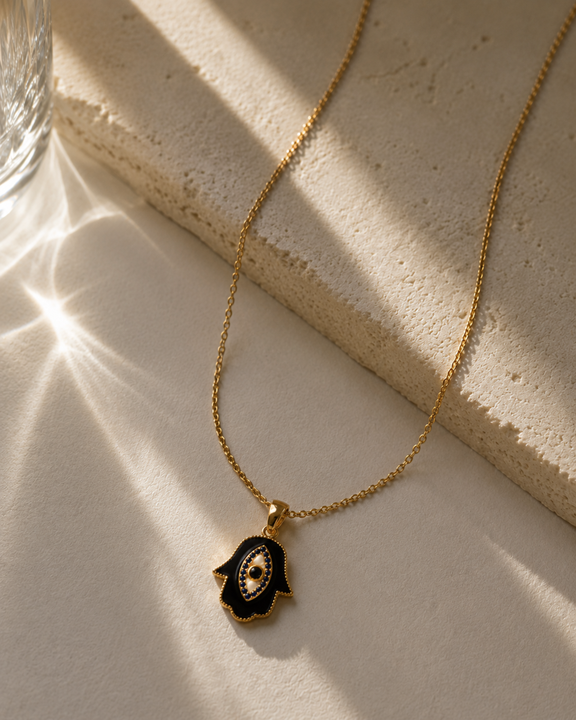 Gold necklace with a black and gold hamsa hand pendant on a beige surface.