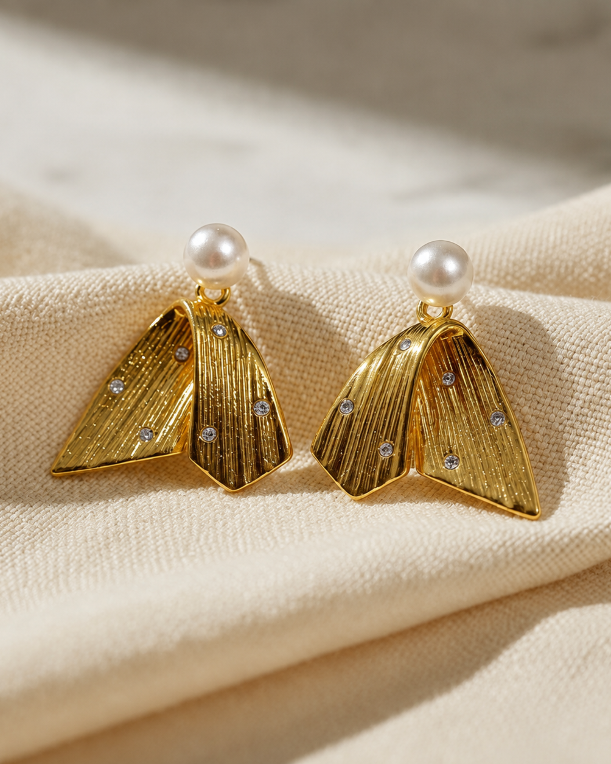 A pair of gold statement earrings with a textured, fan-like geometric design, accented with small clear crystals and topped with white pearl studs, displayed on a beige fabric background.