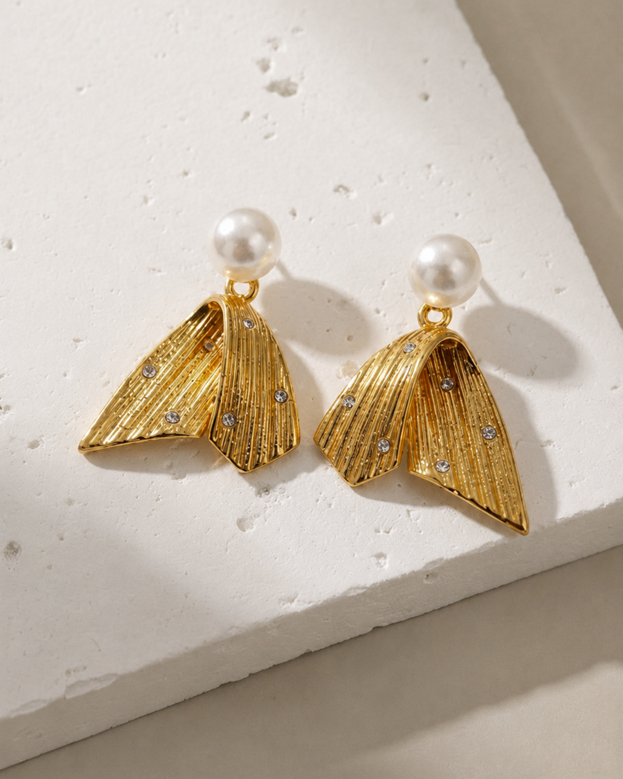 A pair of gold-toned statement earrings with textured, ribbed wing-like shapes and small clear rhinestones, hanging from white faux-pearl studs, displayed on a white textured stone block.