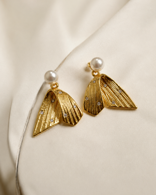 A pair of gold-toned statement earrings with textured leaf-like shapes, small clear rhinestones, and a white pearl at the top, displayed on a soft cream satin fabric.