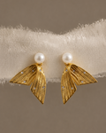 A pair of gold statement earrings with textured leaf-like shapes, a white pearl stud at the top, and small clear rhinestones, displayed on a white fuzzy band against a warm taupe background.