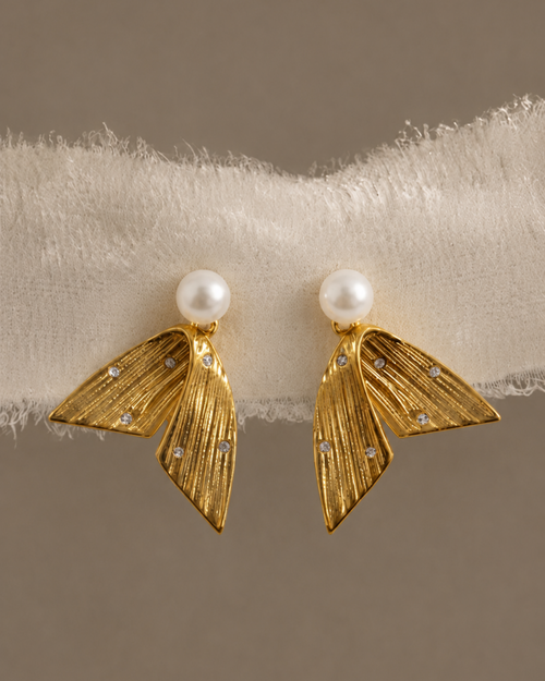 A pair of gold statement earrings with textured leaf-like shapes, a white pearl stud at the top, and small clear rhinestones, displayed on a white fuzzy band against a warm taupe background.