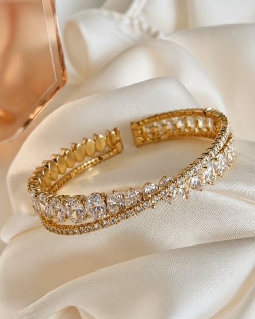 A gold-toned open cuff bracelet embellished with rows of clear crystals, displayed on draped cream satin fabric.