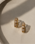 A pair of gold hoop earrings paved with small clear stones, resting on a textured beige surface with soft shadows.