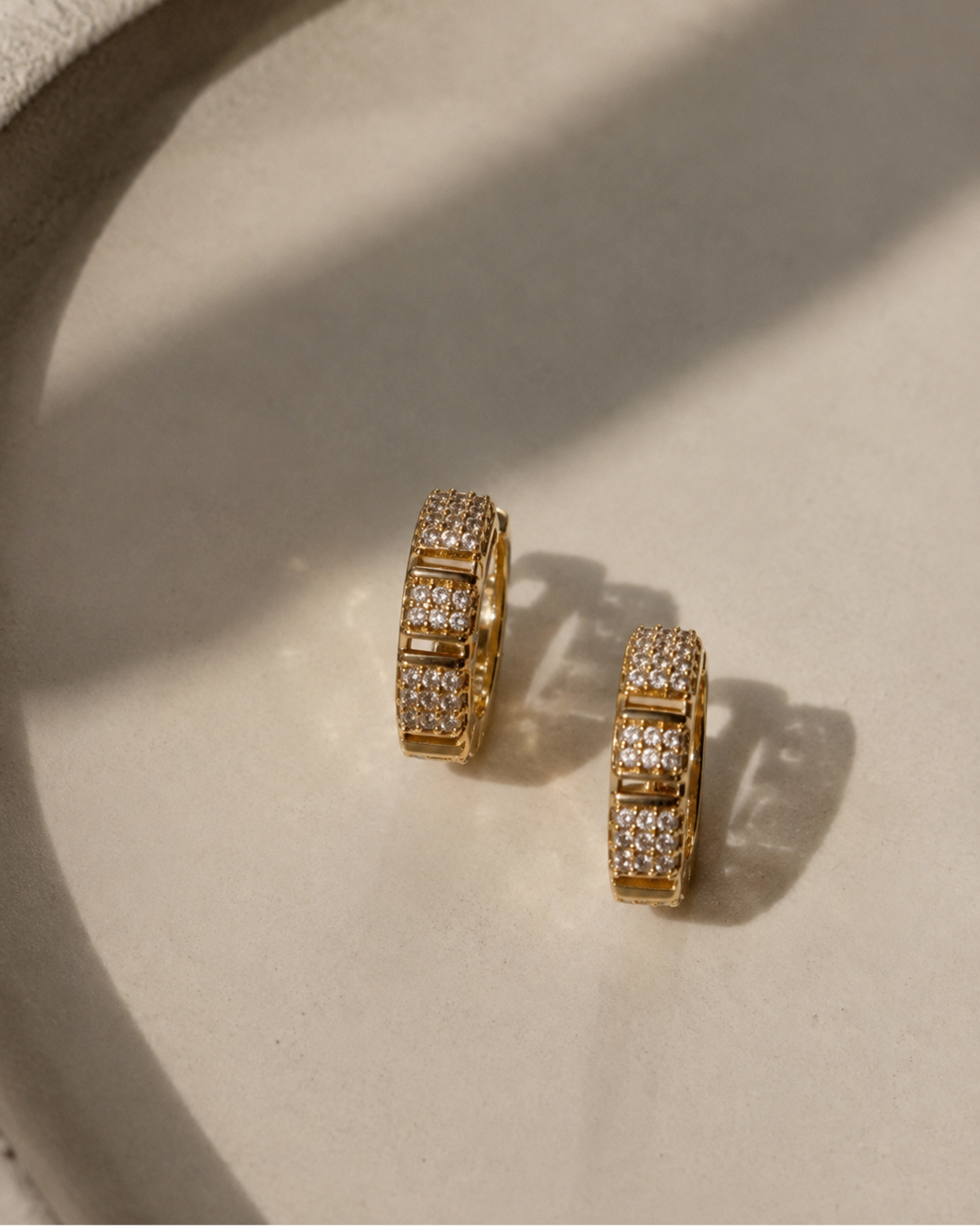 A pair of gold hoop earrings paved with small clear stones, resting on a textured beige surface with soft shadows.