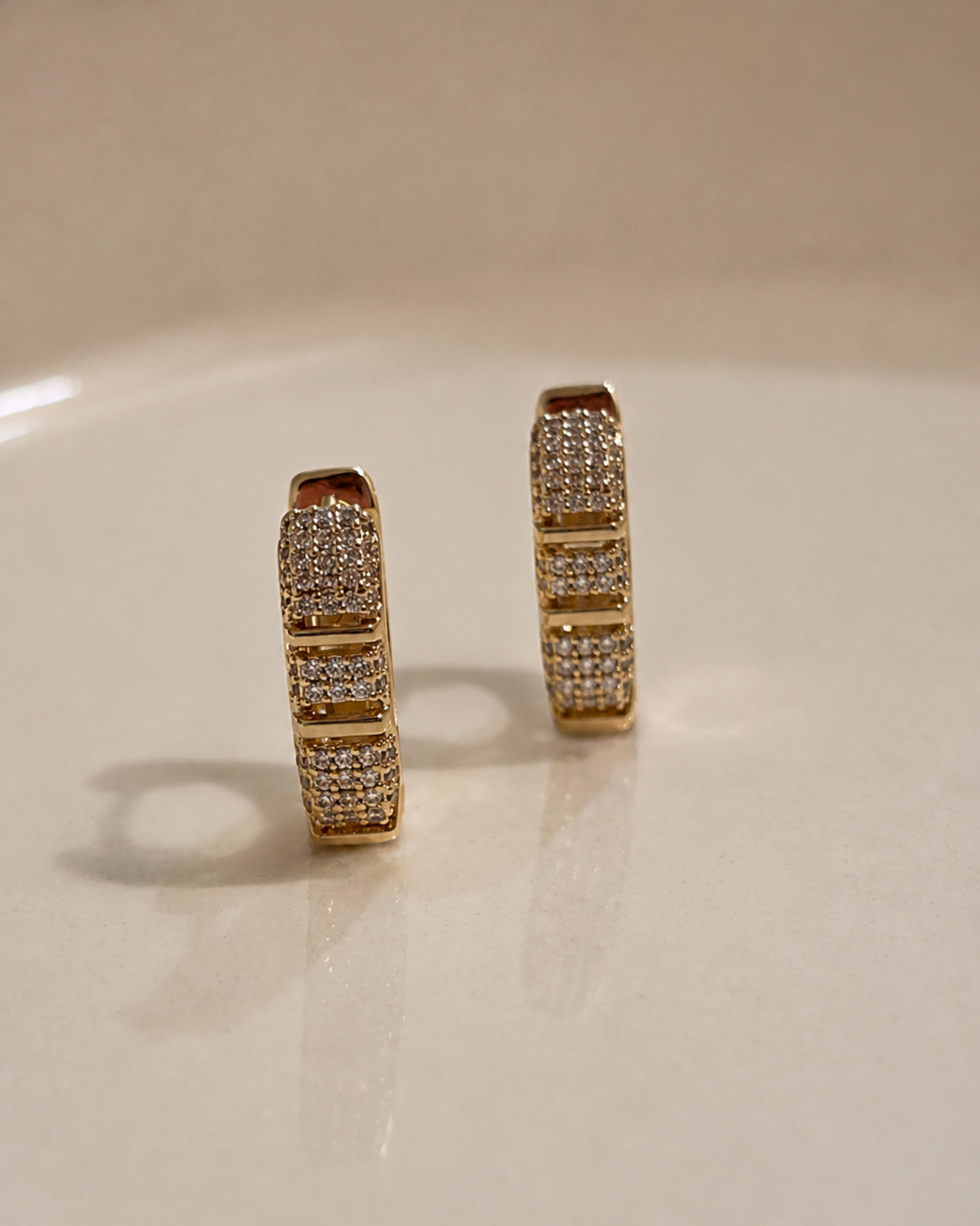 A pair of gold hoop earrings paved with small clear stones, standing upright on a light beige reflective surface.