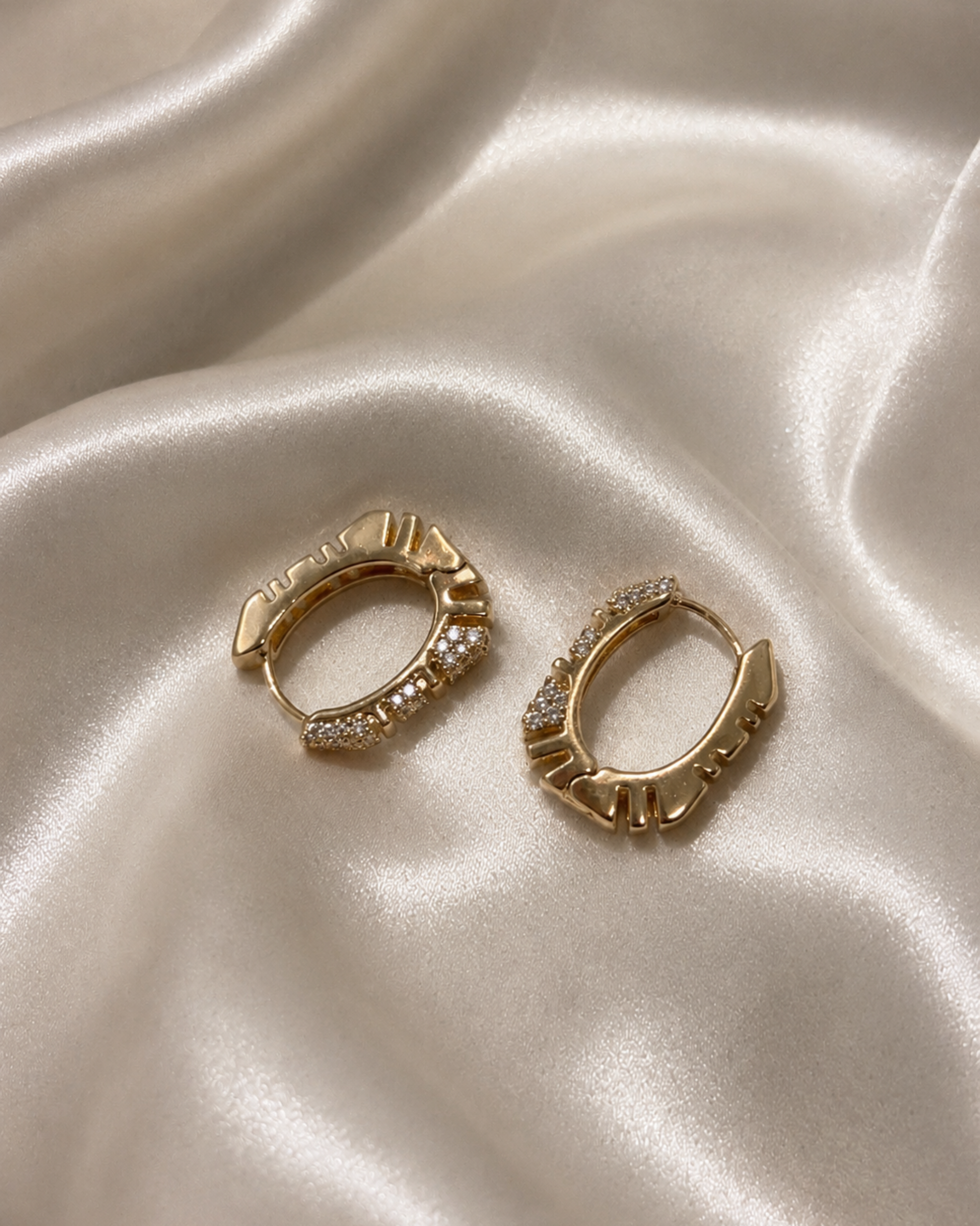 A pair of gold huggie hoop earrings with clear crystal accents, displayed on a draped cream satin fabric background.