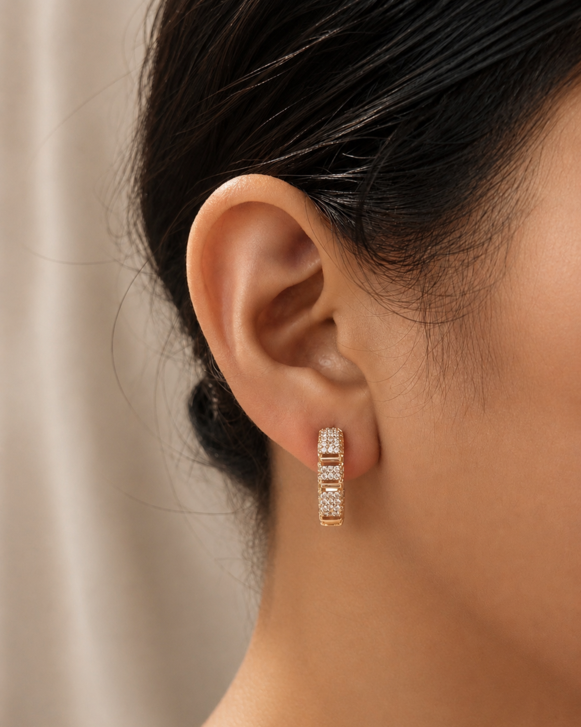A close-up of a model's ear wearing a rose gold huggie hoop earring adorned with small clear pavé-set stones.
