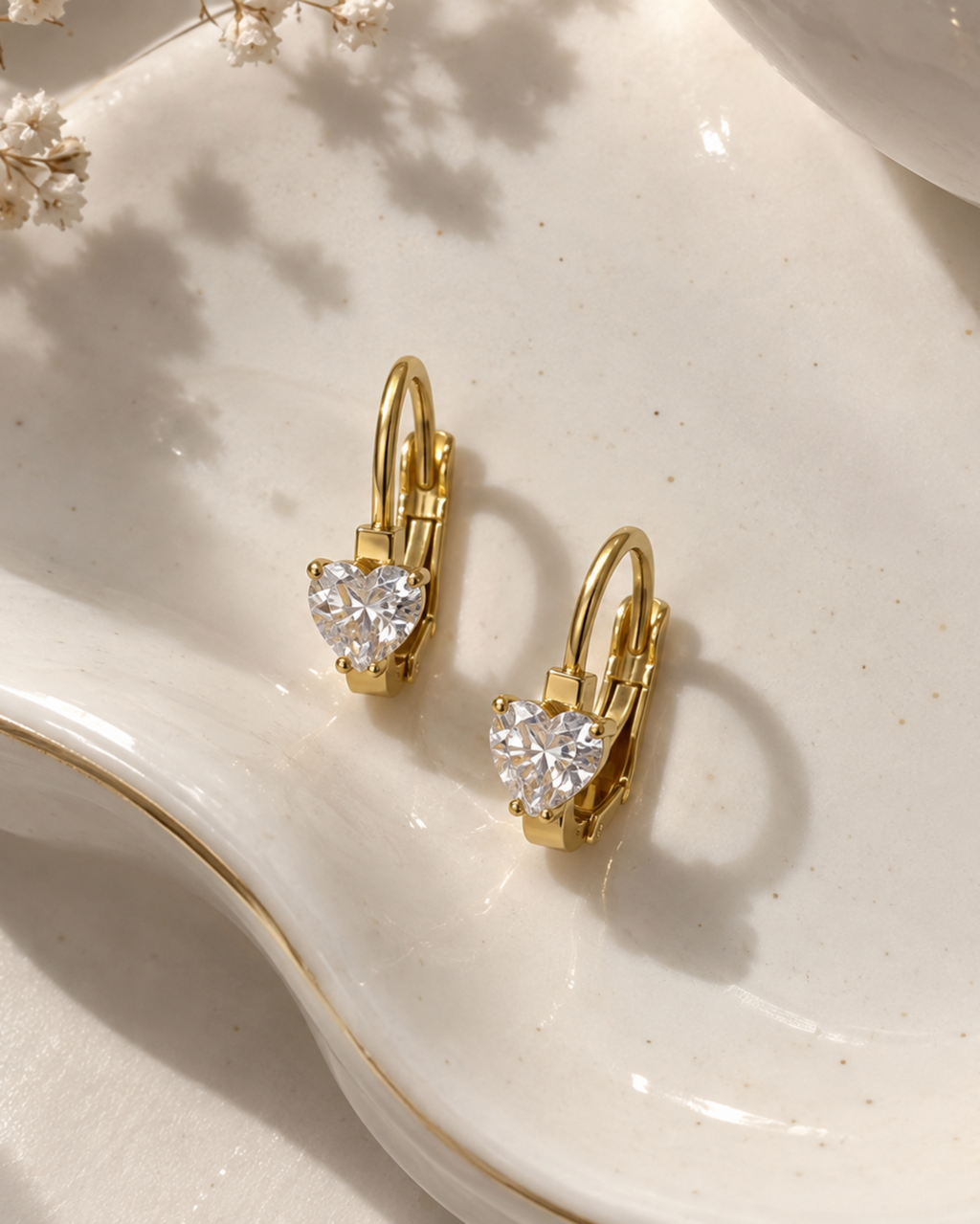 A pair of gold leverback earrings with heart-shaped clear crystal centers, displayed on a white ceramic dish with delicate baby's breath flowers.
