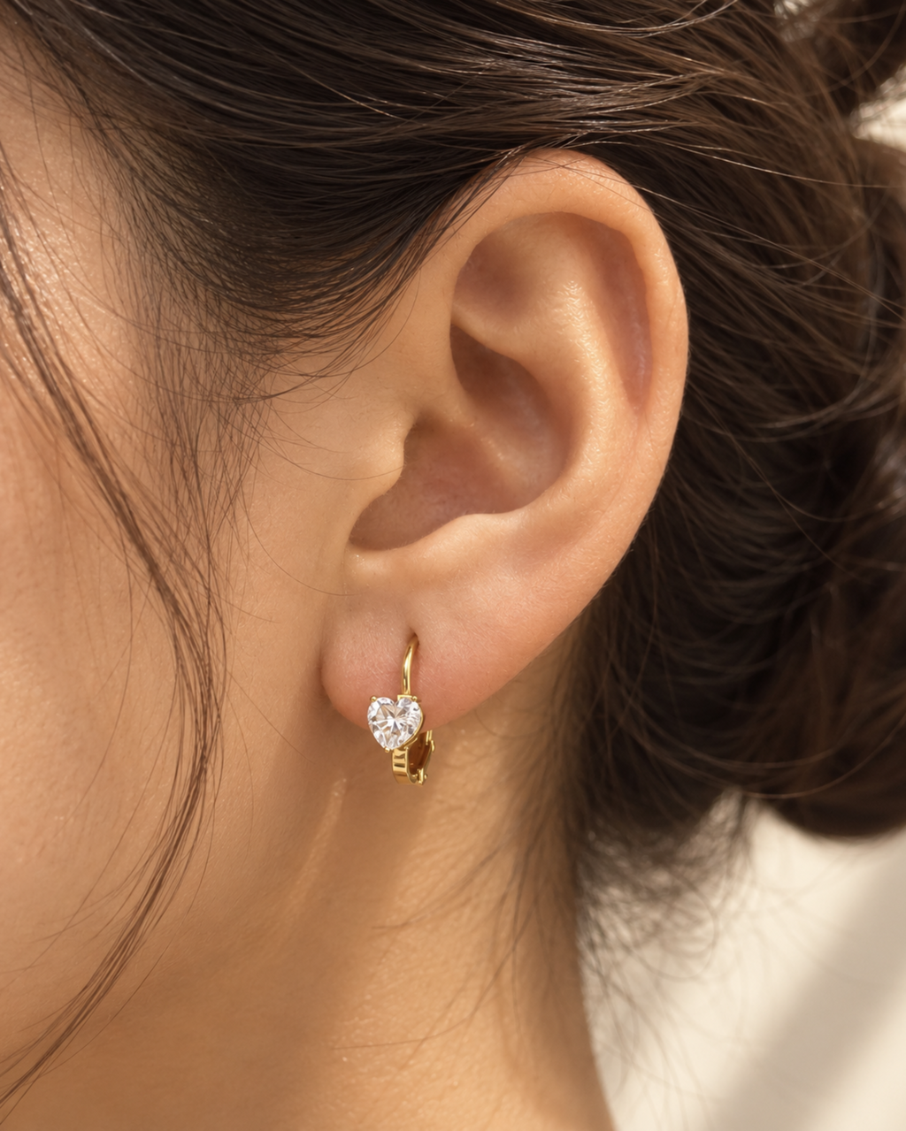 A close-up of a person's ear wearing a small gold huggie hoop earring with a clear heart-shaped crystal.