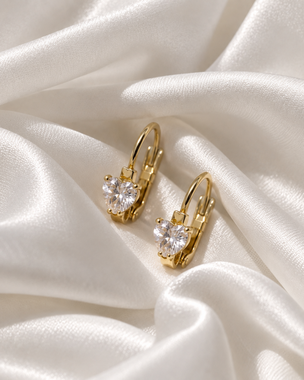 A pair of gold leverback earrings with heart-shaped clear gemstones, displayed on draped white satin fabric.