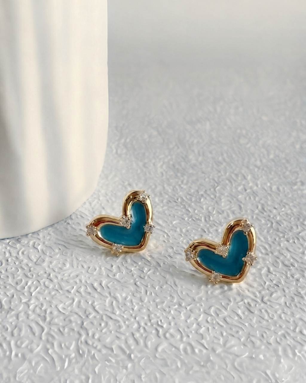 A pair of small heart-shaped stud earrings with turquoise blue centers and gold borders, resting on a textured white surface next to a white ceramic vase.