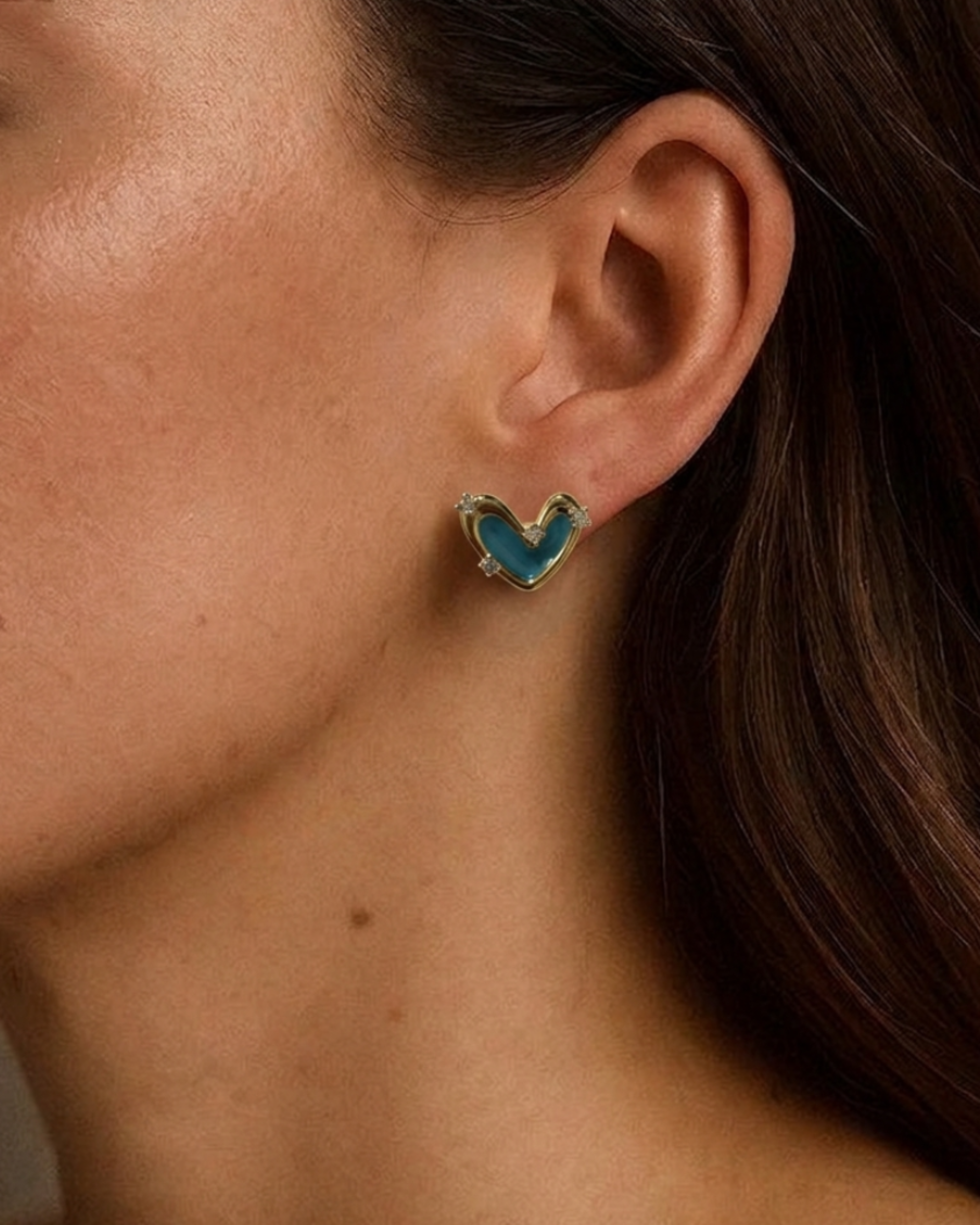 A close-up of a model's ear wearing a small heart-shaped stud earring with a turquoise blue enamel center and gold-tone metal borders.