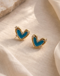 A pair of gold heart-shaped stud earrings with a bright blue enamel center and small clear crystal accents, displayed on draped cream fabric.