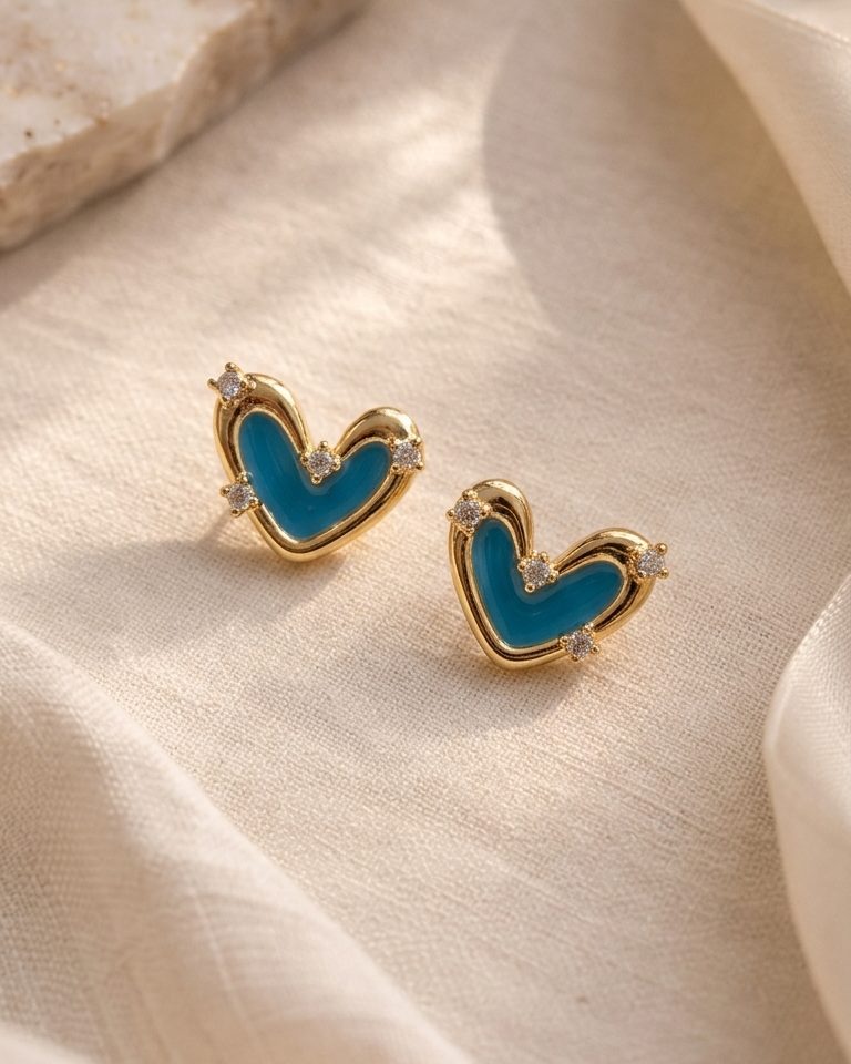 A pair of gold heart-shaped stud earrings with a bright blue enamel center and small clear crystal accents, displayed on draped cream fabric.