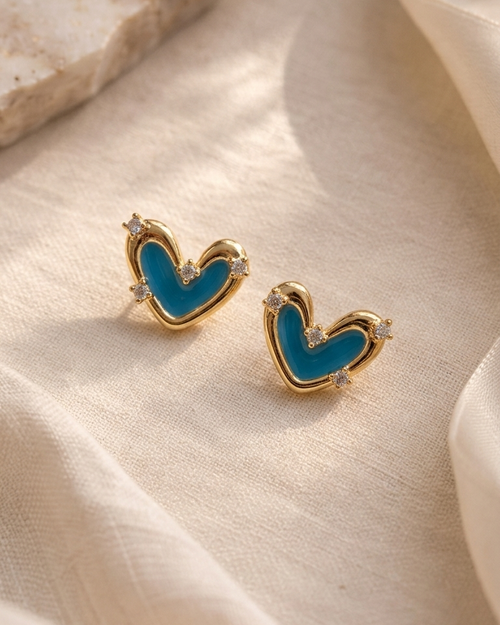 A pair of gold heart-shaped stud earrings with a bright blue enamel center and small clear crystal accents, displayed on draped cream fabric.
