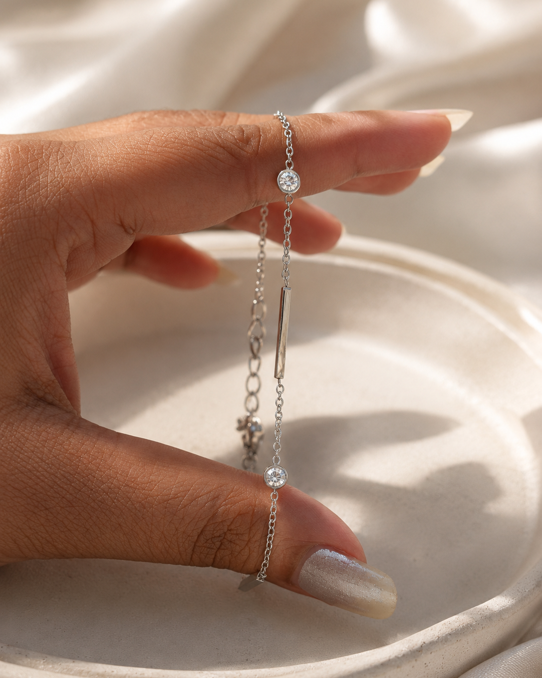 A delicate silver chain bracelet with a central bar, two small round clear stones, and a tiny charm, held between fingers against a soft white satin background.
