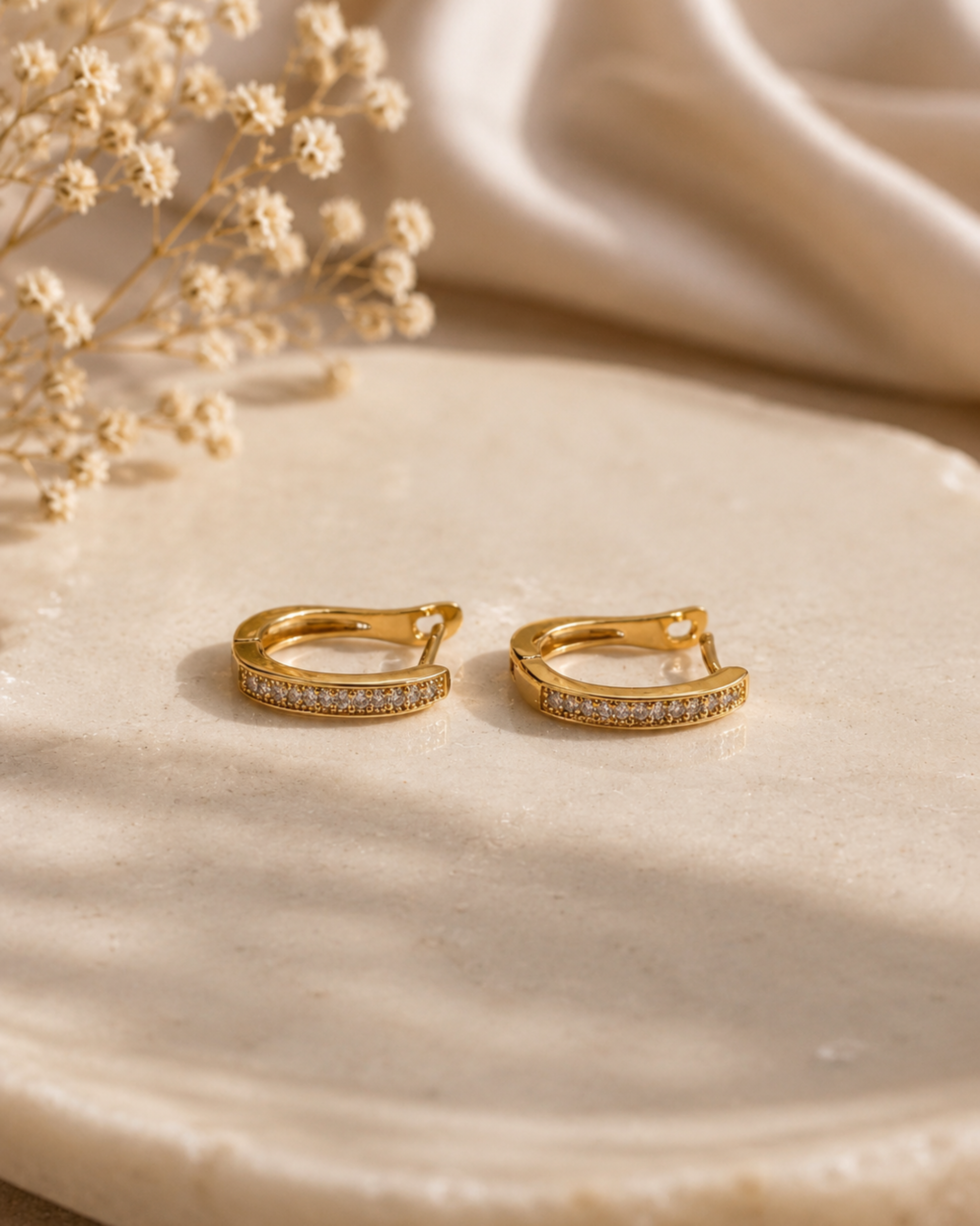 A pair of gold huggie hoop earrings with pavé-set clear stones, displayed on a textured beige stone surface with dried flowers and draped satin fabric in the background.