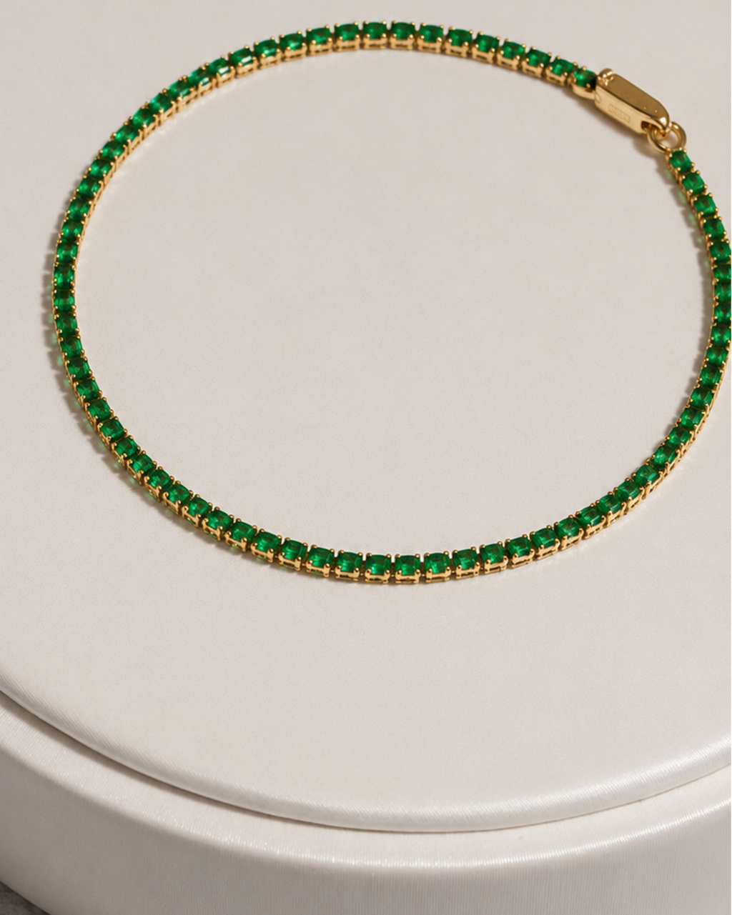 A gold tennis bracelet with a row of small green rectangular stones, displayed on a round white surface.