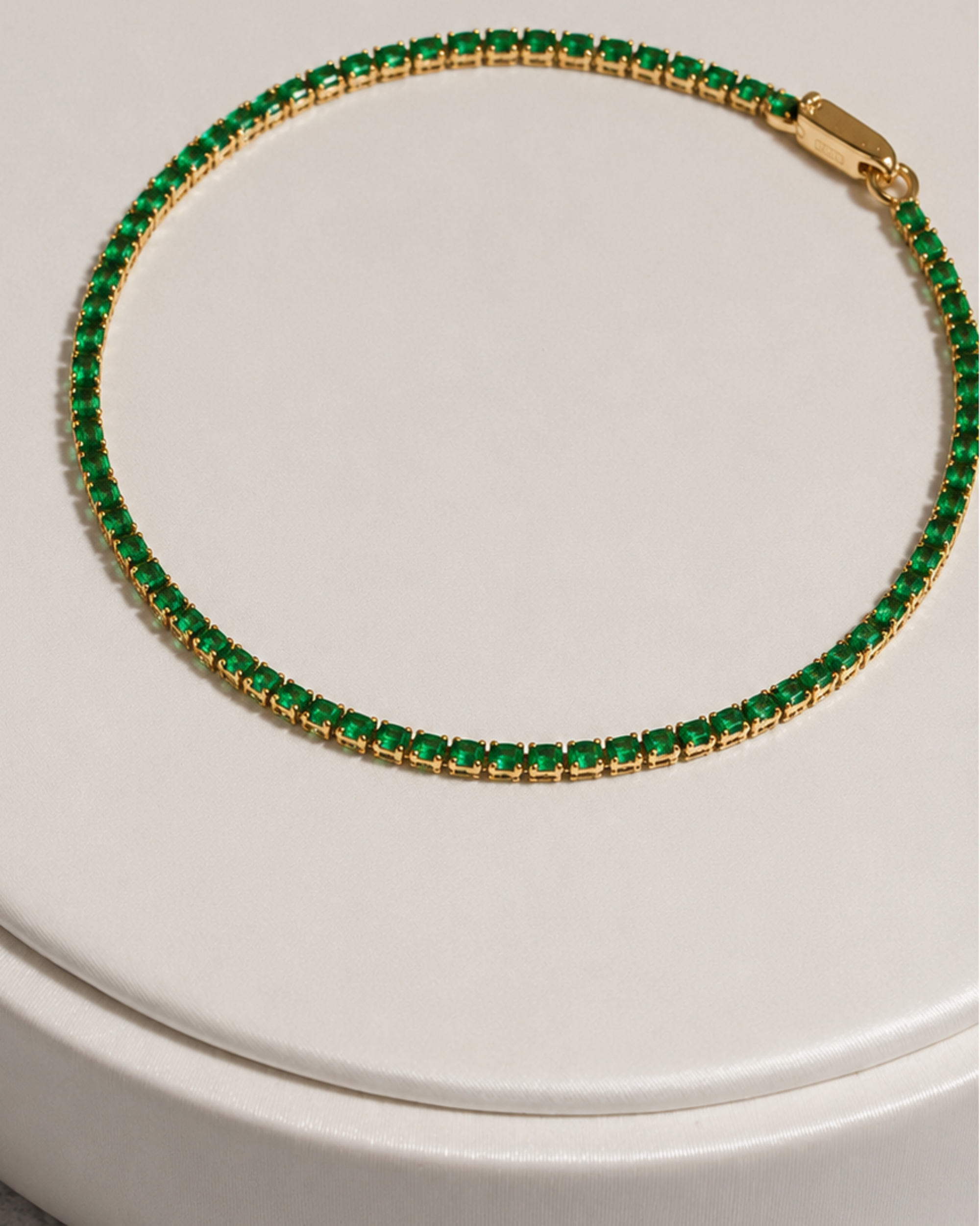 A gold tennis bracelet with a row of small green rectangular stones, displayed on a round white surface.