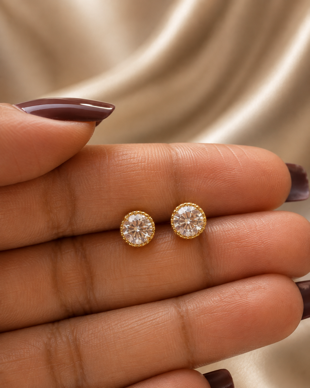 A close-up of a person's hand holding a pair of round gold stud earrings with clear gemstones, set against a soft beige satin background.