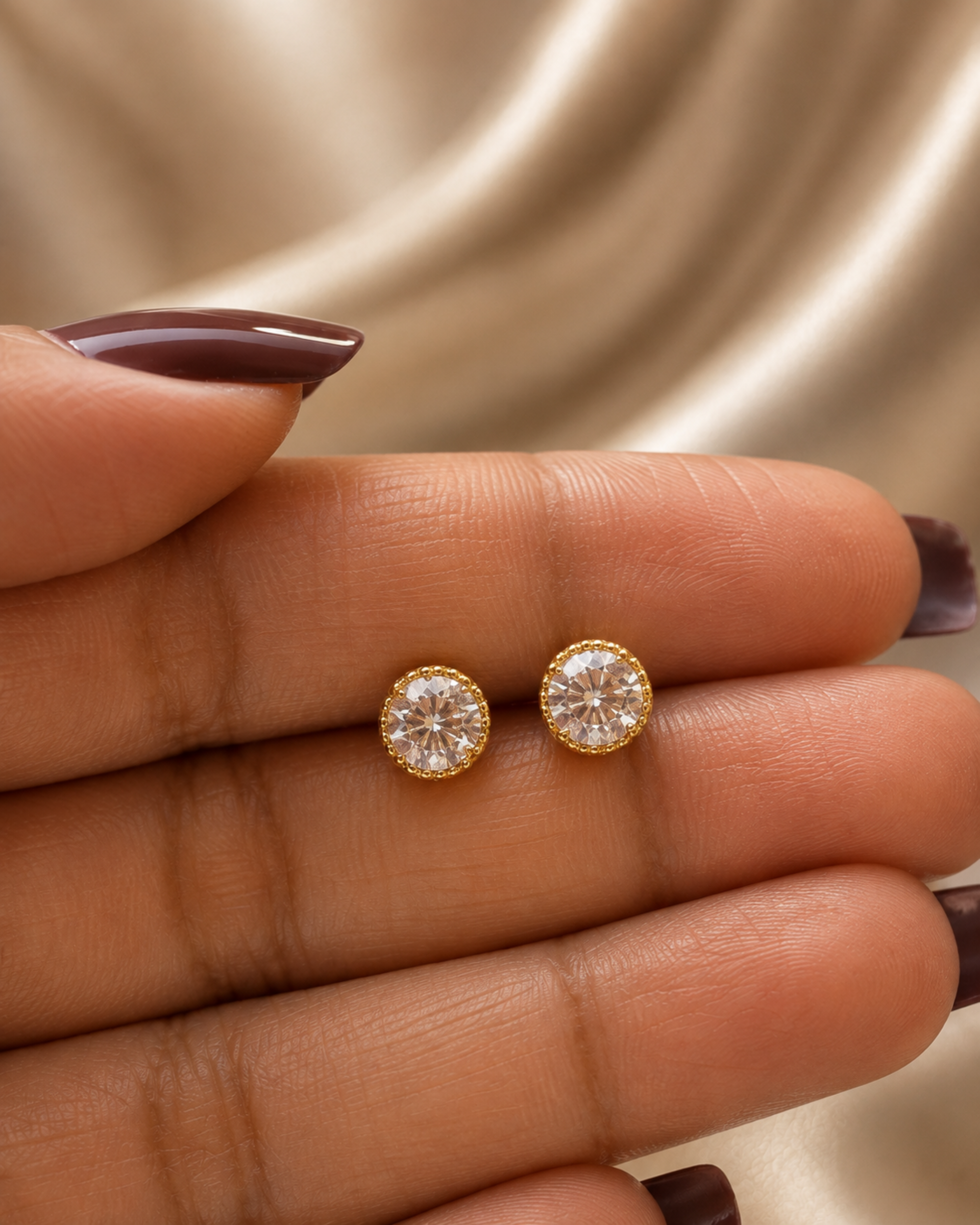 A close-up of a person's hand holding a pair of round gold stud earrings with clear gemstones, set against a soft beige satin background.