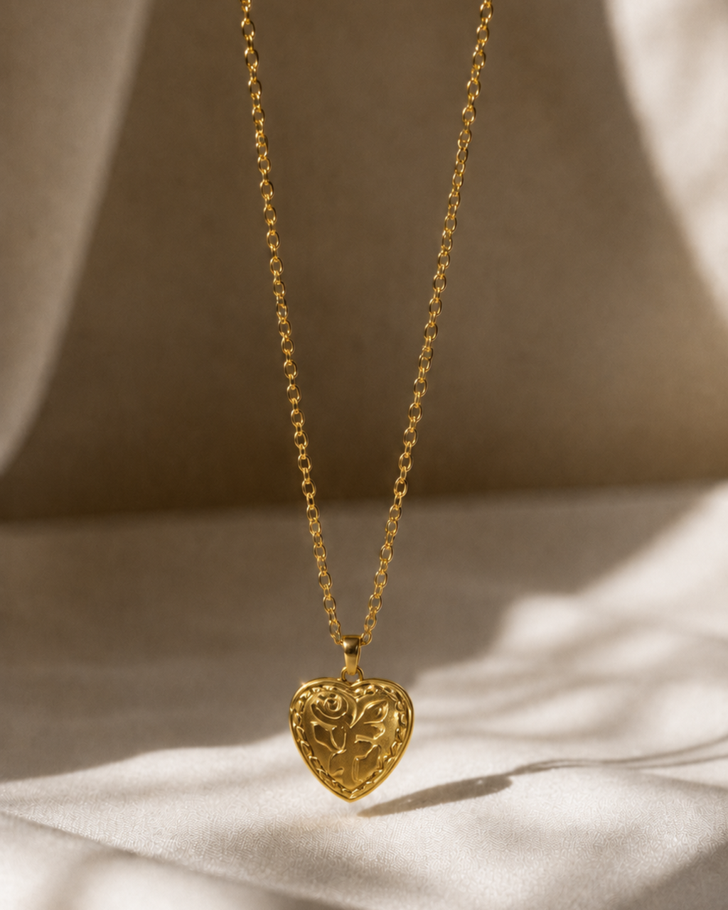 A delicate gold chain necklace with an intricately carved heart-shaped pendant, displayed against a draped beige fabric background with soft, warm shadows.