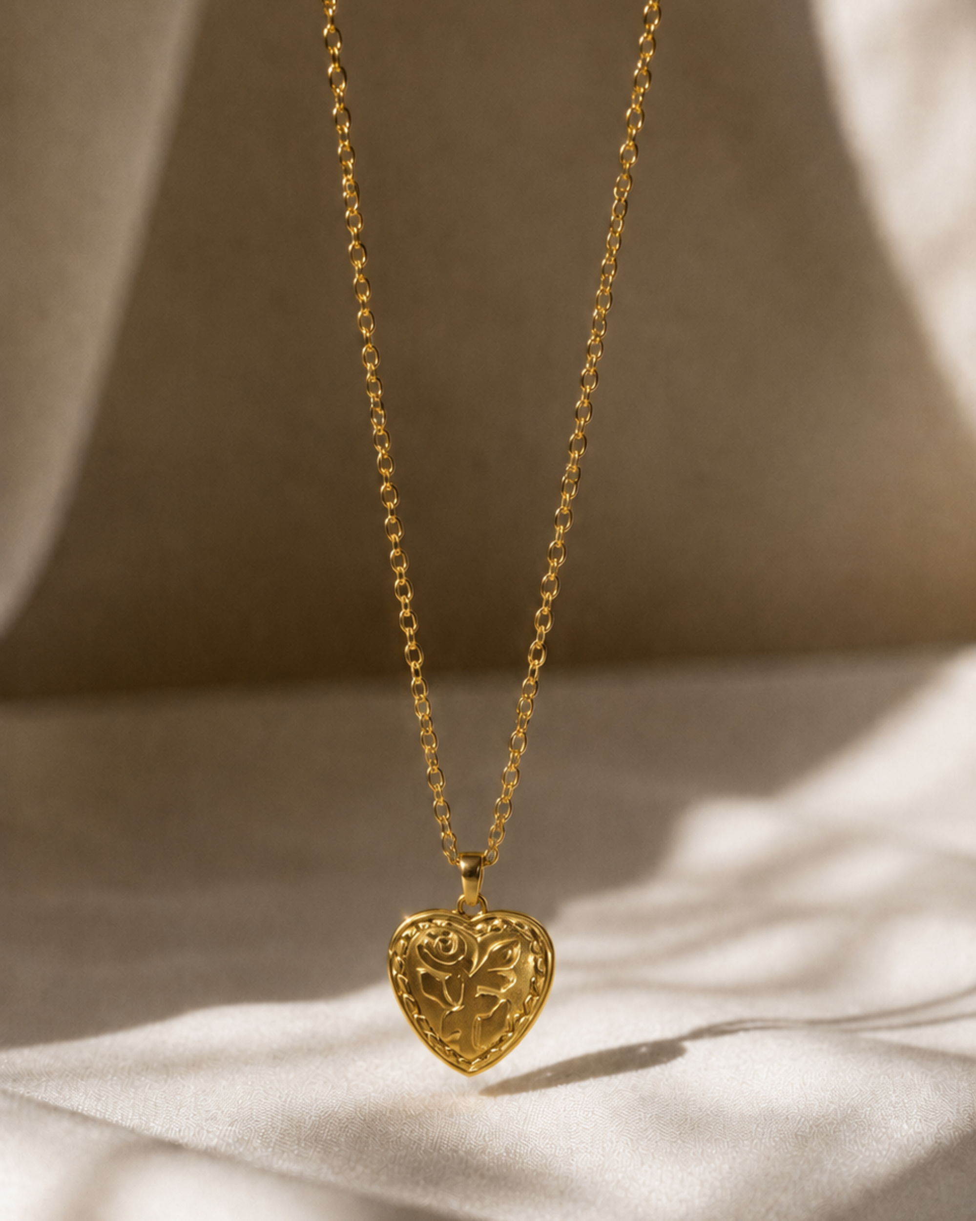 A delicate gold chain necklace with an intricately carved heart-shaped pendant, displayed against a draped beige fabric background with soft, warm shadows.